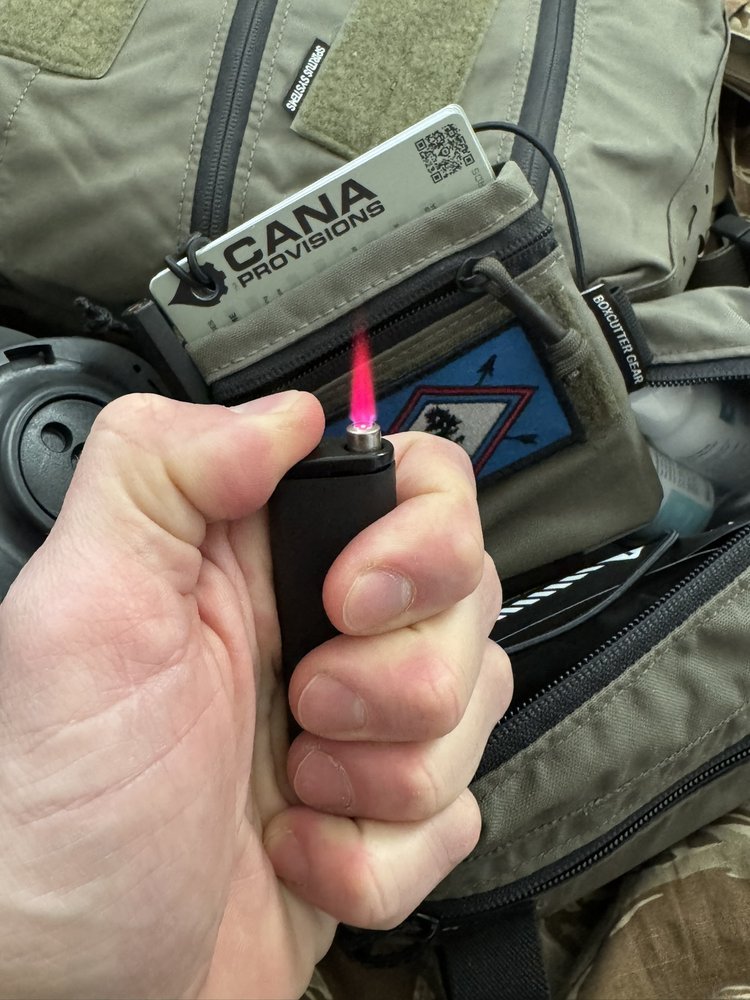 Black CANA MK Torch Pocket Lighter held in hand, lit pink flame, CANA Provisions hang tag on olive tactical gear