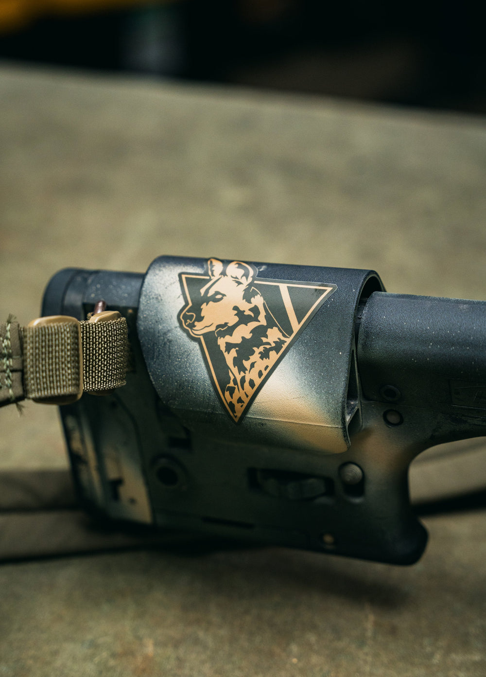 Dirty Civilian die-cut sticker on a matte black rifle stock, tan African Painted Dog and triangle logo, in-use shot