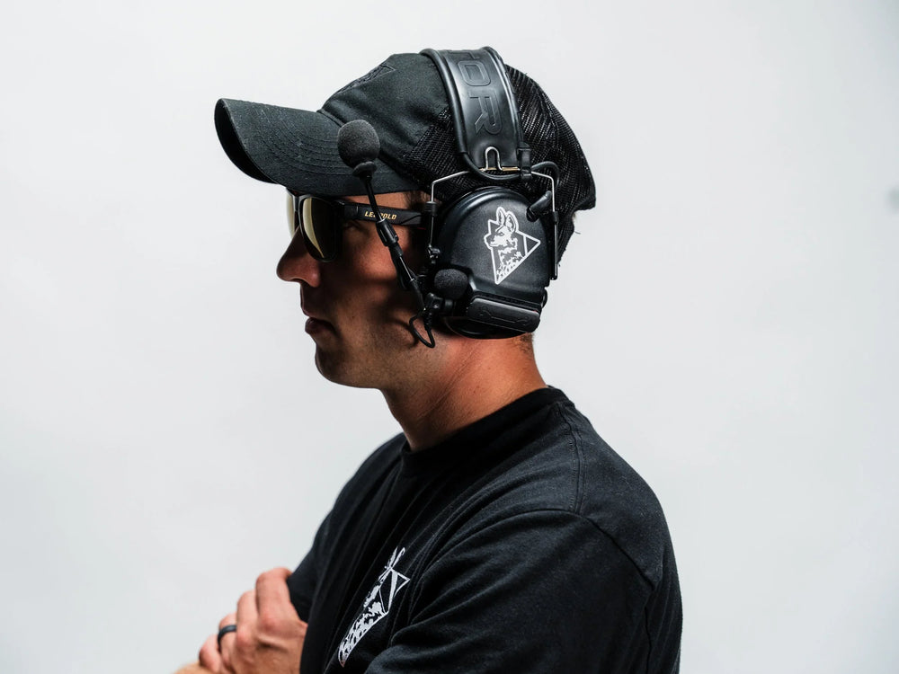 Dirty Civilian painted dog logo sticker applied to black electronic earmuffs, worn in profile by a man in a cap