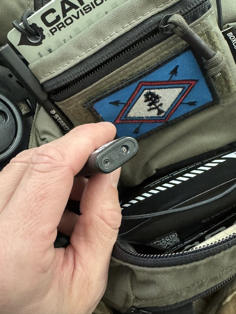 Hand holding the slim gunmetal CANA MK Torch Pocket Lighter, top-end view showing twin burner ports, olive gear behind