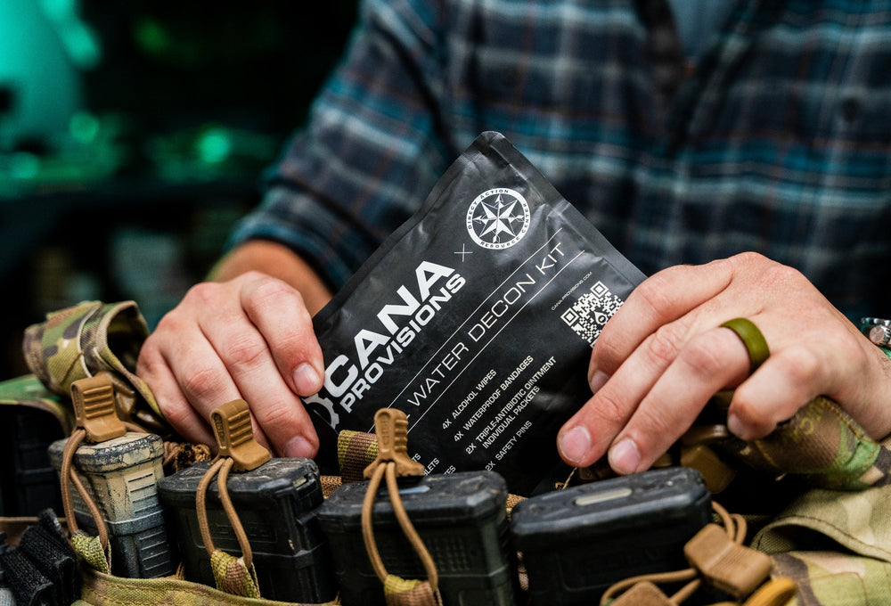 Hands tucking the black CANA Provisions Water Decon Kit pouch into camo chest-rig mag pouches, in-use shot